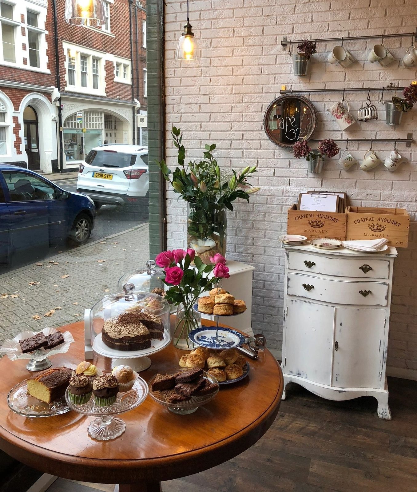 Cakes and bakes on a round wooden table by the window at Café 32, with teacups hanging on the white brick wall