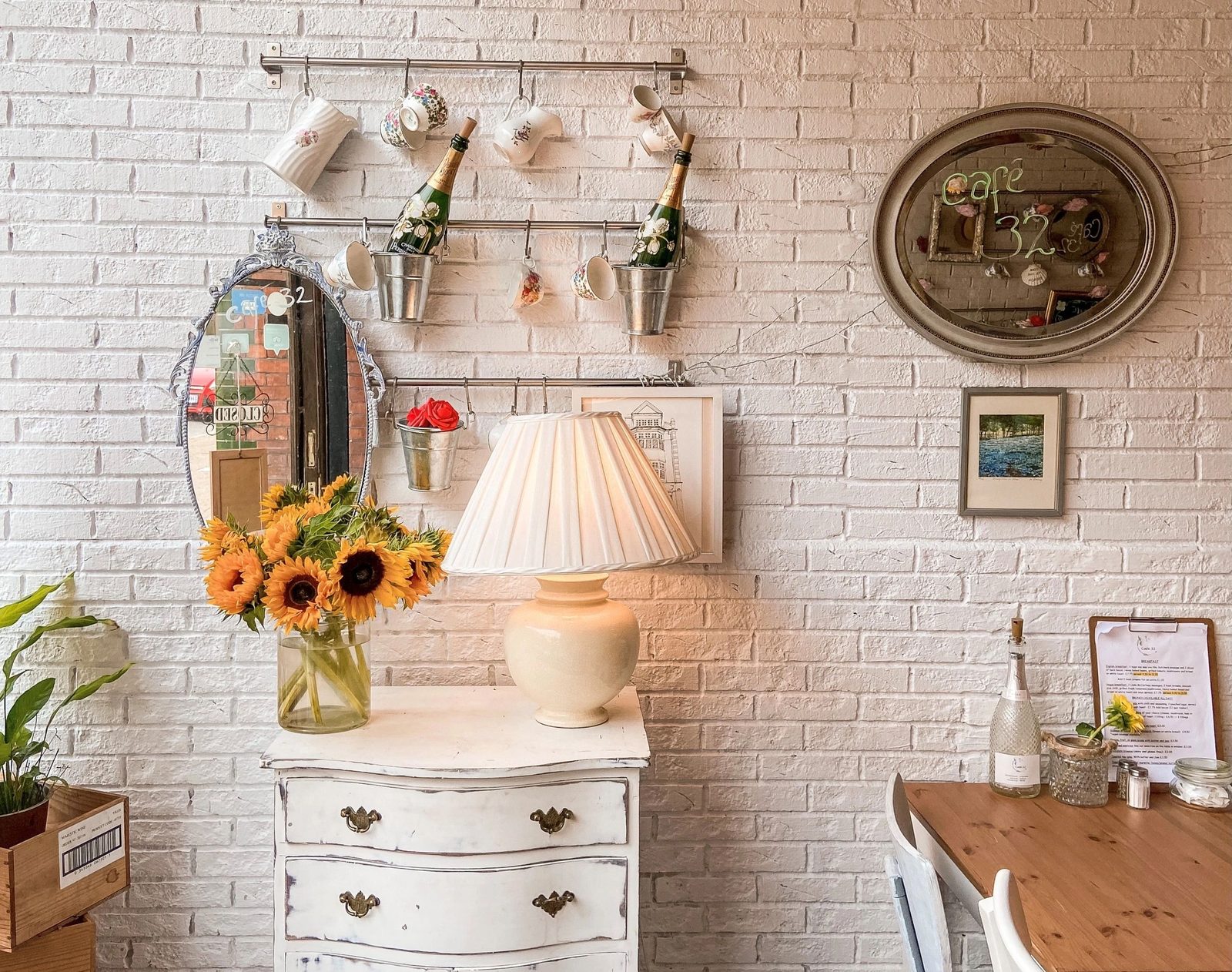 Inside Café 32: white-painted brick, sunflowers in a jug, a lamp on a vintage white chest of drawers and an oval mirror
